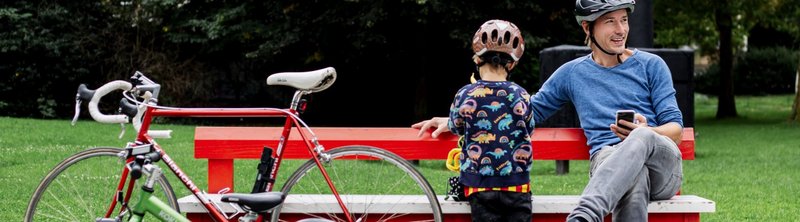 Mann und Kind sitzen auf einer roten Bankim Park, daneben steht ein Fahrrad.